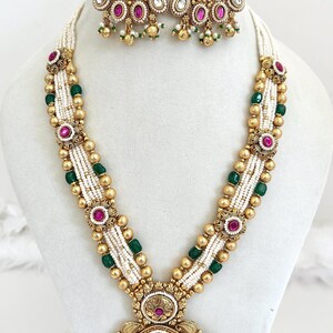 May include: An ornate gold-toned necklace and matching earrings set. The necklace features white beaded strands, green and gold accents, and a large pendant with a green center. The earrings have a similar design with red and white accents.