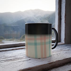 Könnte beinhalten: Eine schwarze Kaffeetasse mit pastellfarbenem Karomuster und schwarzem Henkel. Die Tasse ist mit einem heißen Getränk gefüllt, wie der aufsteigende Dampf zeigt. Die Tasse steht auf einer verwitterten, weiß gestrichenen Holzoberfläche.