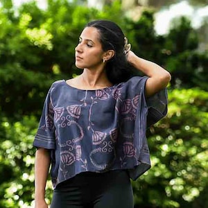 May include: A dark blue top with a floral pattern in shades of pink and white. The top has short sleeves and a loose fit. The model is wearing black leggings. The background is a lush green garden.