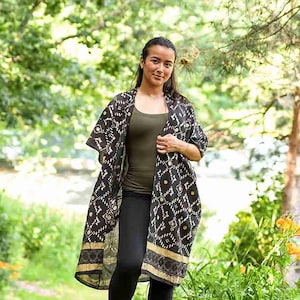 May include: A woman wearing a black and gold patterned kimono-style jacket over a green top and black leggings. The jacket has a geometric design and a gold border. The background is a lush green outdoor setting with trees and foliage.
