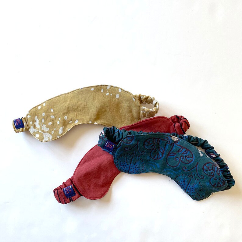May include: Three sleep masks in the image. One is tan with white spots, another is burgundy, and the third is teal with a floral pattern. Each mask has an elastic band and a small blue tag.