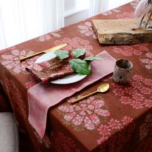 May include: A brown tablecloth with a pink and green floral pattern. The tablecloth is set with a white plate, a gold fork and spoon, a pink napkin, and a brown ceramic cup.