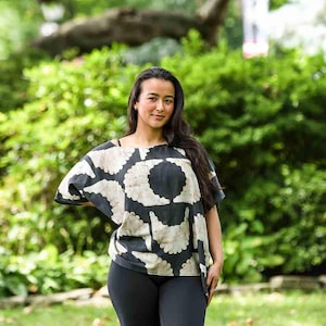 Könnte beinhalten: Eine Person trägt ein Oberteil mit schwarz-weißem Muster und schwarze Leggings vor einem grünen, belaubten Hintergrund. Das Oberteil hat ein sich wiederholendes kreisförmiges Design in Schwarz und Off-White. Die Leggings sind figurbetont.