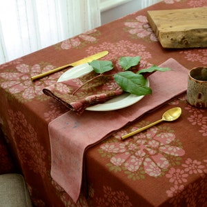 May include: A rust-coloured tablecloth with a floral pattern in shades of pink and green. The tablecloth is set with a white plate, a pink napkin, a gold fork and knife, and a gold spoon. A sprig of green leaves is placed on the plate.