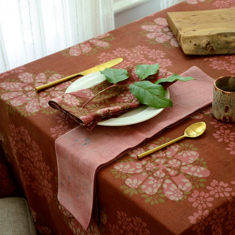 May include: A rust-coloured tablecloth with a floral pattern in shades of pink and green. The tablecloth is set with a white plate, a pink napkin, a gold fork and knife, and a gold spoon. A sprig of green leaves is placed on the plate.