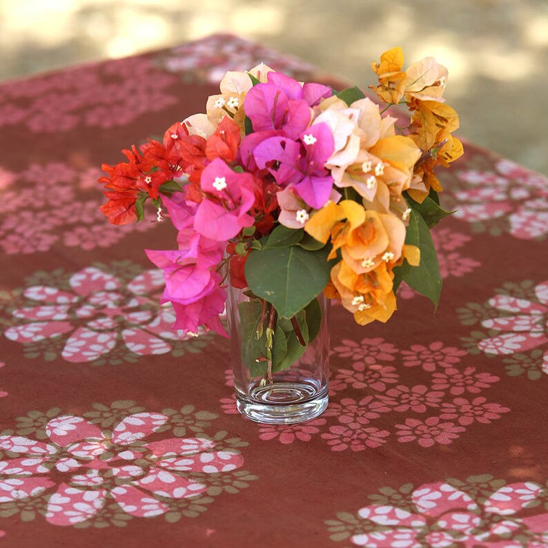 May include: A bouquet of pink, orange and red bougainvillea flowers in a clear glass vase on a brown table with a white floral pattern.