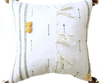 May include: White cushion with grey stripes and white fringe. The cushion has yellow pom-poms on the corners and sides.