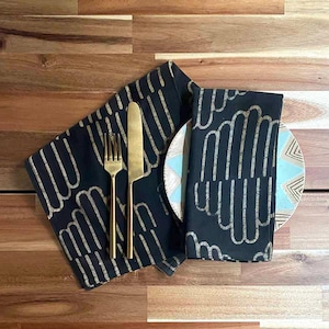 May include: Two black napkins with a gold geometric pattern are folded on a wooden table. A gold fork and knife are placed on top of the napkins. A blue and white patterned plate is also on the table.