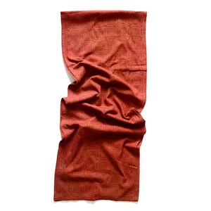 May include: A folded, rectangular scarf in a reddish-brown hue. The fabric features a repeating geometric pattern in a slightly lighter shade of the same color. The scarf is draped, showing folds and texture.