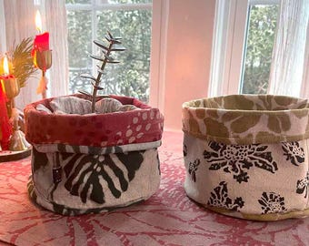 May include: Two cylindrical fabric baskets with folded rims. The left basket features a red patterned top rim and a black leaf print on a cream base. The right basket has a mustard yellow floral pattern on the rim and black floral motifs on cream.