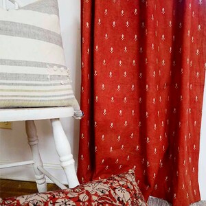 May include: A red curtain with a white floral pattern hangs in a room. A white chair with a striped pillow sits in front of the curtain. A pillow with a floral pattern sits on the floor in front of the curtain.