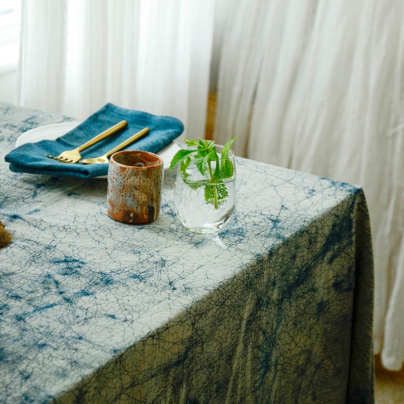 May include: A blue and white patterned tablecloth with a brown ceramic mug, a glass of water with a sprig of green leaves, and a blue napkin with gold silverware.