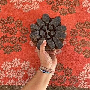 May include: A wooden block printing stamp with a floral design. The stamp is brown and has a detailed pattern of leaves and flowers. The stamp is being held by a hand against a red fabric background with a white floral pattern.
