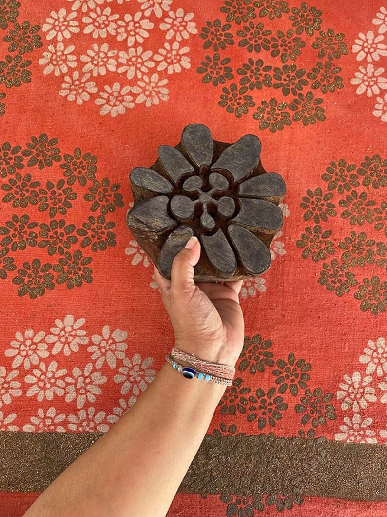 May include: A wooden block printing stamp with a floral design. The stamp is brown and has a detailed pattern of leaves and flowers. The stamp is being held by a hand against a red fabric background with a white floral pattern.
