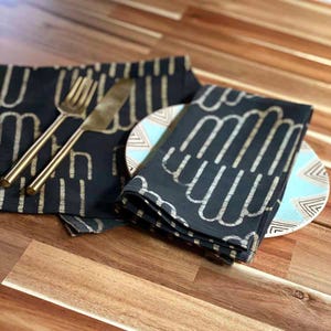 May include: Black and gold patterned fabric napkins with a geometric design. The napkins are folded and placed on a wooden surface with a white and blue patterned plate.