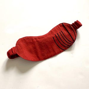 May include: Red sleep mask with black curved lines printed on the front. The mask has an elastic band.