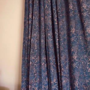 Peut inclure: Rideau bleu marine avec un motif floral en rose pâle. Le tissu est drapé, montrant des plis verticaux. Le motif présente des fleurs stylisées et des formes de feuilles, créant un aspect texturé. Le rideau est suspendu à côté d'un mur uni blanc cassé.