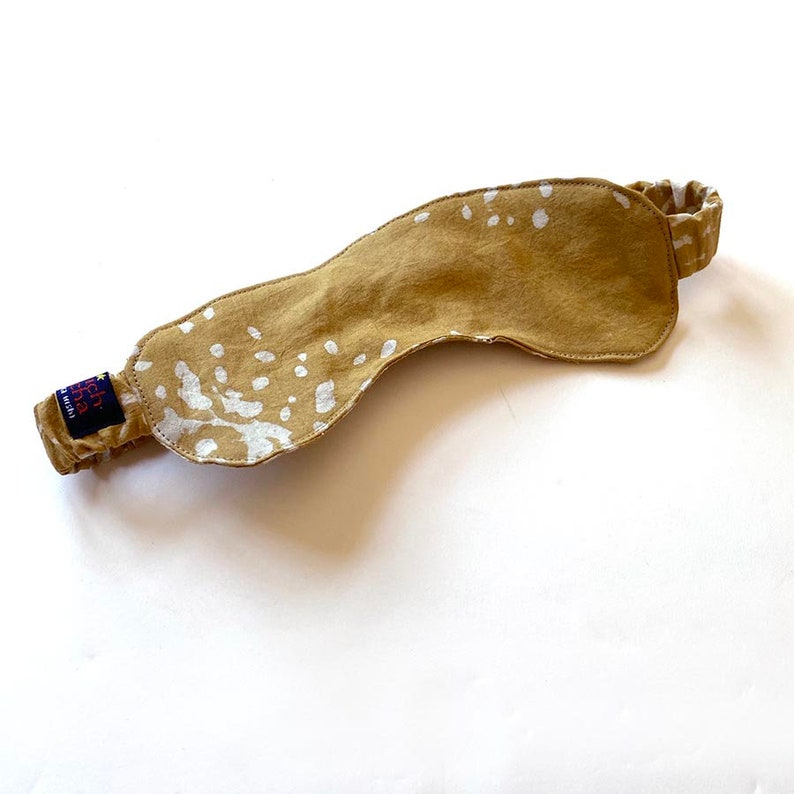 May include: A tan sleep mask with a white splatter pattern. The mask has an elastic band for a comfortable fit.