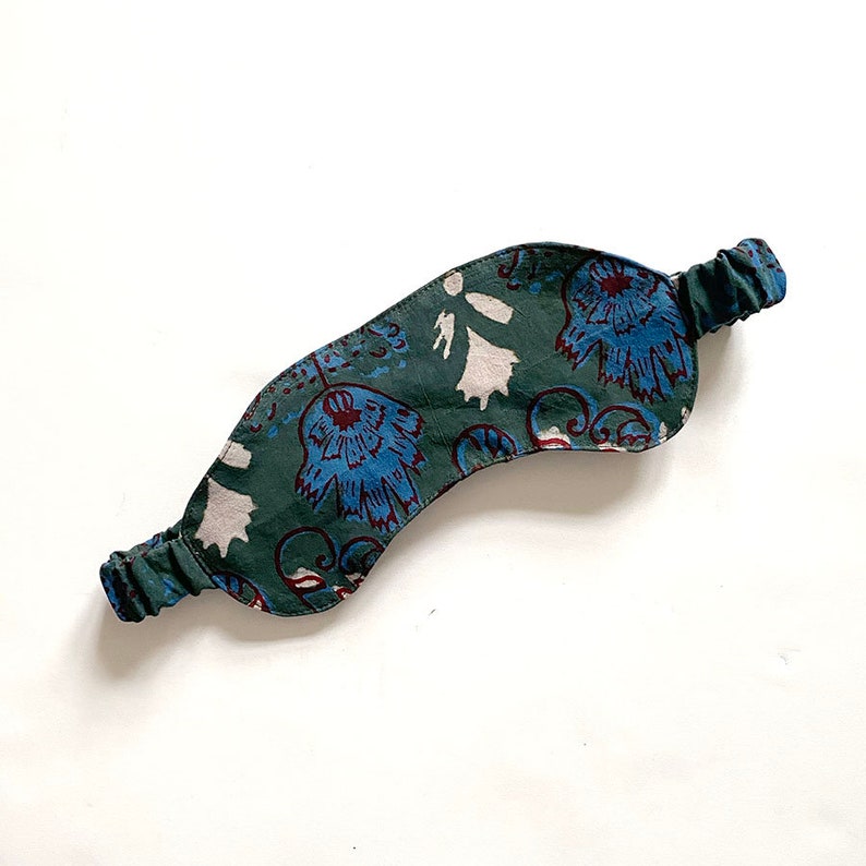 May include: A green sleep mask with a floral pattern in blue and white. The mask has an elastic band for a comfortable fit. The design features stylized flowers and swirling patterns, creating a calming aesthetic. The mask is designed to block out light for better sleep.