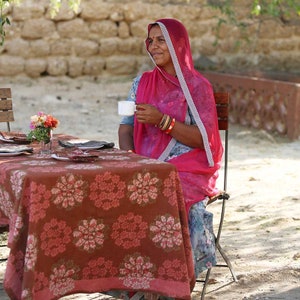 May include: A woman wearing a pink and white patterned sari sits at a table with a floral tablecloth. She is holding a white cup and looking off to the side.