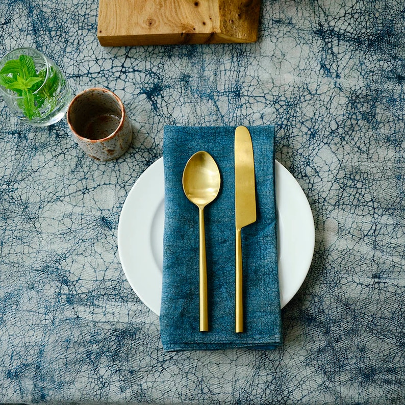 May include: A blue linen napkin with a gold knife and spoon on a white plate. The plate is on a blue and white patterned tablecloth.