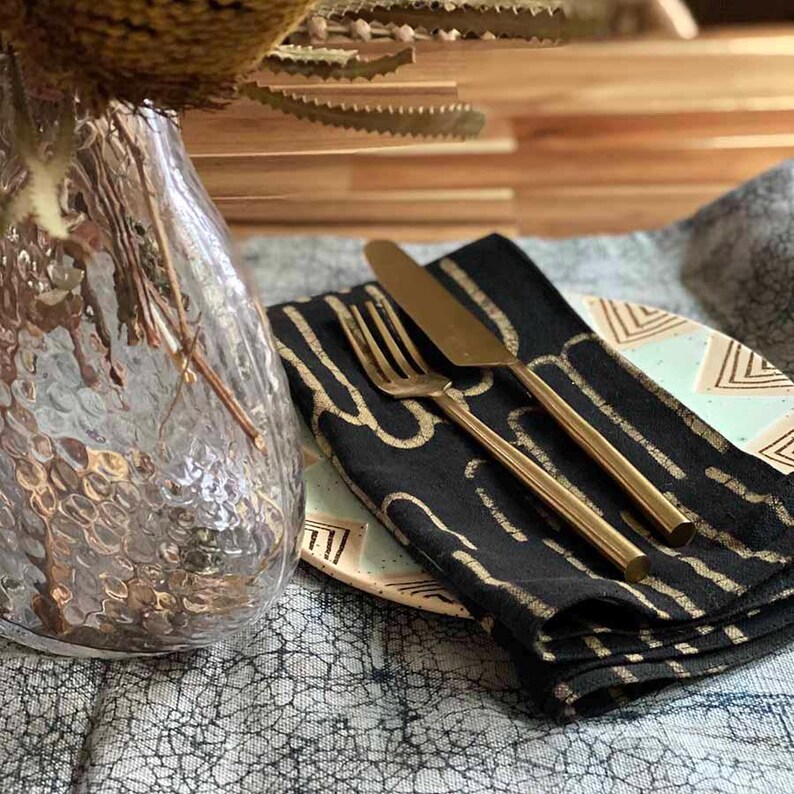 May include: Black linen napkins with gold geometric patterns, folded on a light blue and white patterned plate with a gold fork and knife.