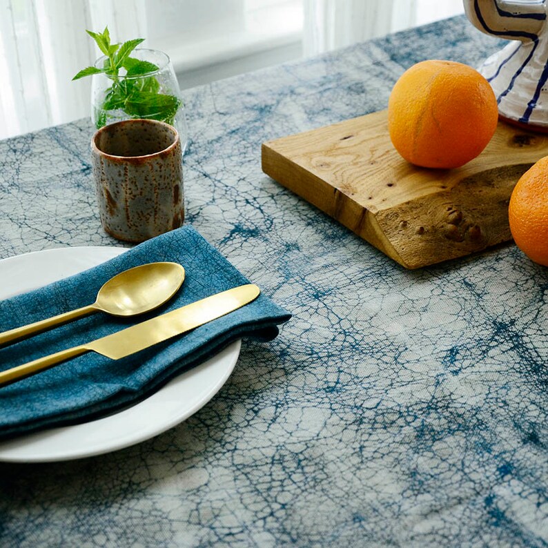 May include: A blue and white patterned tablecloth with a white plate, gold silverware, and a blue napkin. There is a wooden cutting board with two oranges on it.