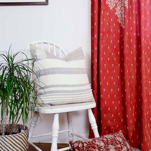 May include: A white wooden chair with a striped pillow and a red patterned throw pillow. A potted plant is in the foreground. The chair is in front of a red patterned curtain.