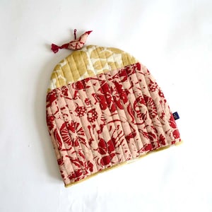 May include: A quilted tea cozy with a floral pattern in red and cream. The cozy has a bird-shaped ornament attached to the top. The cozy is made of fabric and has a gold trim.