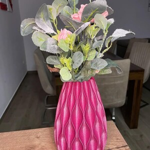 May include: A bright magenta vase with a vertical, repeating wave-like textured pattern. The vase holds an arrangement of artificial greenery featuring frosted, sage-green leaves and small, light pink flower buds at the top.