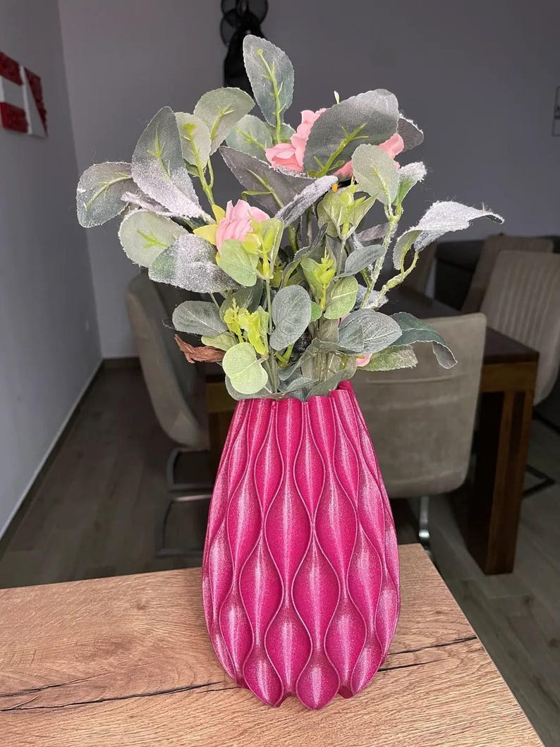 May include: A bright magenta vase with a vertical, repeating wave-like textured pattern. The vase holds an arrangement of artificial greenery featuring frosted, sage-green leaves and small, light pink flower buds at the top.
