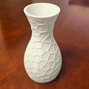 May include: A light gray vase with a textured, geometric pattern. The vase has a wide base that tapers to a narrower neck and flares out at the top. The surface has a speckled appearance.