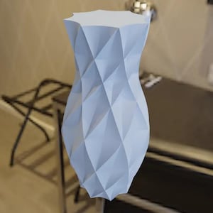 Op de afbeelding: Een lichtblauwe decoratieve vaas met een geometrisch, diamantachtig patroon. De vaas heeft een brede, facetgeslepen bovenkant en een gebogen, zandloperachtige vorm. De vaas is gemaakt van een solide materiaal en heeft een matte afwerking.