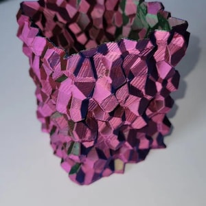 Op de afbeelding: Een 3D-geprinte vaas met een geometrisch ontwerp in roze, paarse en groene tinten. Het oppervlak heeft een textuur die bestaat uit talrijke polygonale vormen. Dit decoratieve item kan worden gebruikt als bloempot of als uniek woonaccessoire.