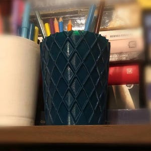 Op de afbeelding: Een tealkleurige pennenhouder met een ruitpatroon, gevuld met kleurrijke pennen en potloden. Links staat een witte keramische beker gevuld met pennen. De achtergrond is een boekenplank.