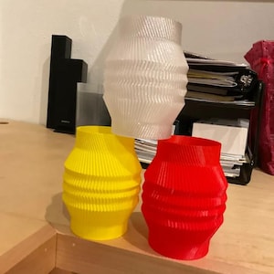 Puede incluir: Una colección de jarrones decorativos en varios colores. Los jarrones tienen un diseño único y acanalado y se muestran en blanco, amarillo y rojo. Los jarrones están hechos de un material translúcido y están colocados sobre una superficie de madera.