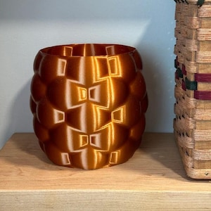 Pode incluir: Um vaso de cor cobre, impresso em 3D, com um padrão geométrico texturizado. O vaso cilíndrico tem uma abertura larga e assenta numa prateleira de madeira. Uma cesta tecida com detalhes verdes e bordô é parcialmente visível.