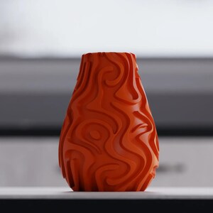 Op de afbeelding: Een levendige oranje vaas met een uniek, wervelend patroon in het oppervlak. De vaas heeft een afgeronde vorm en een brede opening, geschikt voor het uitstallen van bloemen of als decoratief stuk. Het ontwerp heeft vloeiende, organische lijnen.