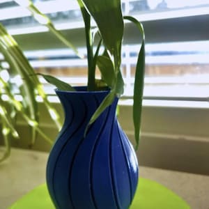 Op de afbeelding: Een blauwe vaas met een uniek, gebogen ontwerp, met groene bladplanten. De vaas staat op een felgroen oppervlak, met een raam en jaloezieën op de achtergrond. De vaas is ongeveer 15 cm hoog.