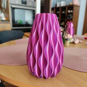 May include: A vibrant magenta vase with a repeating vertical wave pattern. The surface features a glossy, iridescent finish that catches the light, creating deep shadows and bright highlights along the curved, textured ridges of the cylindrical form.