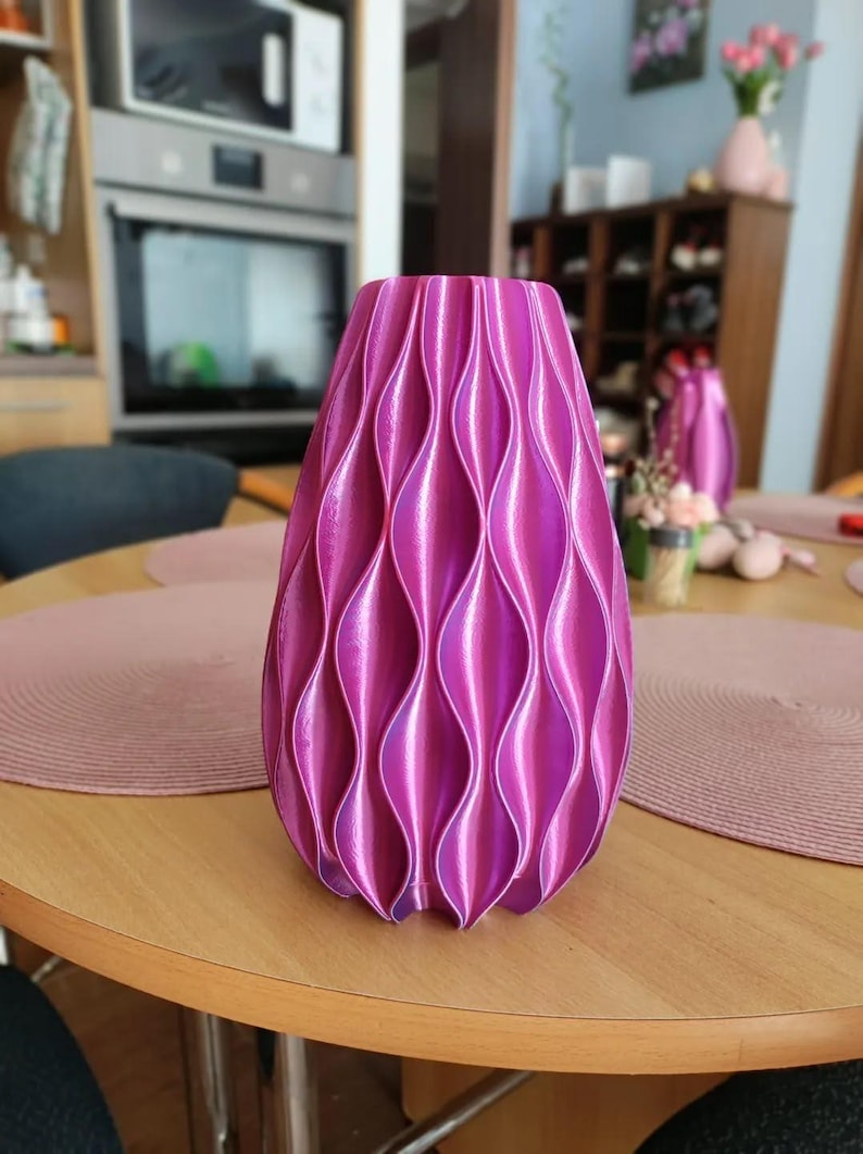 May include: A vibrant magenta vase with a repeating vertical wave pattern. The surface features a glossy, iridescent finish that catches the light, creating deep shadows and bright highlights along the curved, textured ridges of the cylindrical form.