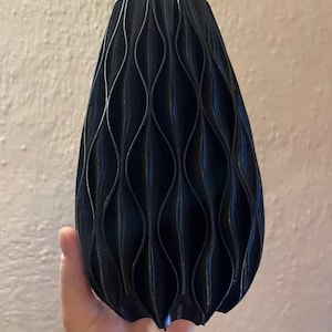 May include: A black vase with a repeating vertical wave pattern across its exterior surface. The object has a tapered shape that is wider at the base and narrows towards the top, featuring a series of deep, curved ridges that create a textured look.