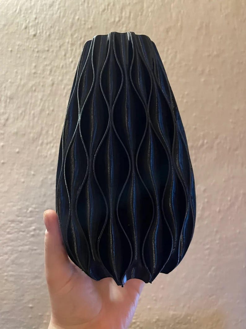 May include: A black vase with a repeating vertical wave pattern across its exterior surface. The object has a tapered shape that is wider at the base and narrows towards the top, featuring a series of deep, curved ridges that create a textured look.