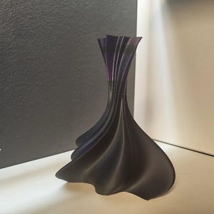 Può includere: Un vaso decorativo viola scuro con un design unico e fluido. Il vaso ha una base ampia che si assottiglia verso l'alto in una serie di elementi verticali curvi. Il vaso è realizzato in un materiale liscio e poggia su una superficie bianca.