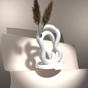 May include: A contemporary white ceramic vase with a unique, looping form. The vase contains two tall, dried brown ornamental grasses. The vase rests on a circular base, set against a neutral beige backdrop.