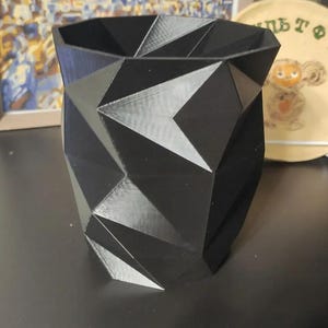 Op de afbeelding: Een zwarte geometrische plantenbak met een facetontwerp. De plantenbak heeft een brede opening en een reeks hoekige, driehoekige vormen. De plantenbak is gemaakt van een solide, ondoorzichtig materiaal.