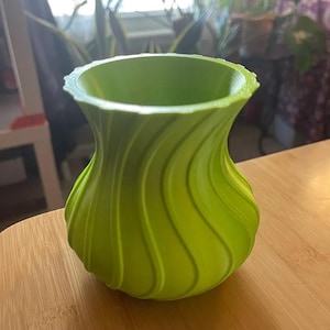 Op de afbeelding: Een heldergroene vaas met een uniek, wervelend ontwerp. De vaas heeft een brede, afgeronde basis die smaller wordt naar de hals en aan de bovenkant uitloopt. Het oppervlak heeft verticale, gebogen ribbels, waardoor het een textuur krijgt. De vaas staat op een houten oppervlak.