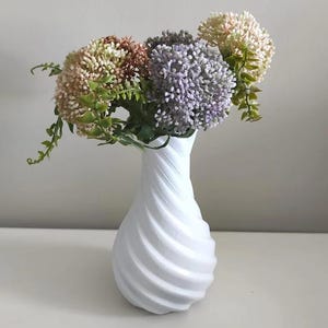 Op de afbeelding: Een witte vaas met spiraaltextuur bevat een boeket kunstbloemen. De bloemen zijn een mix van kleuren, waaronder beige, bruin, paars en groen. De vaas heeft een modern design en staat op een wit oppervlak.