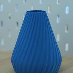 Op de afbeelding: Een levendige blauwe vaas met een uniek, getextureerd spiraalontwerp. De vaas heeft een brede basis die taps toeloopt naar een smallere opening. De vaas is gemaakt van een stevig materiaal en staat op een houten oppervlak.