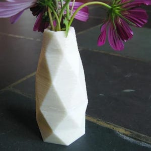 Op de afbeelding: Een witte, geometrische vaas met kosmosbloemen met paarse bloemblaadjes. De vaas heeft een facetontwerp en is gemaakt van een solide, ondoorzichtig materiaal. De bloemen staan in volle bloei en geven de compositie kleur.